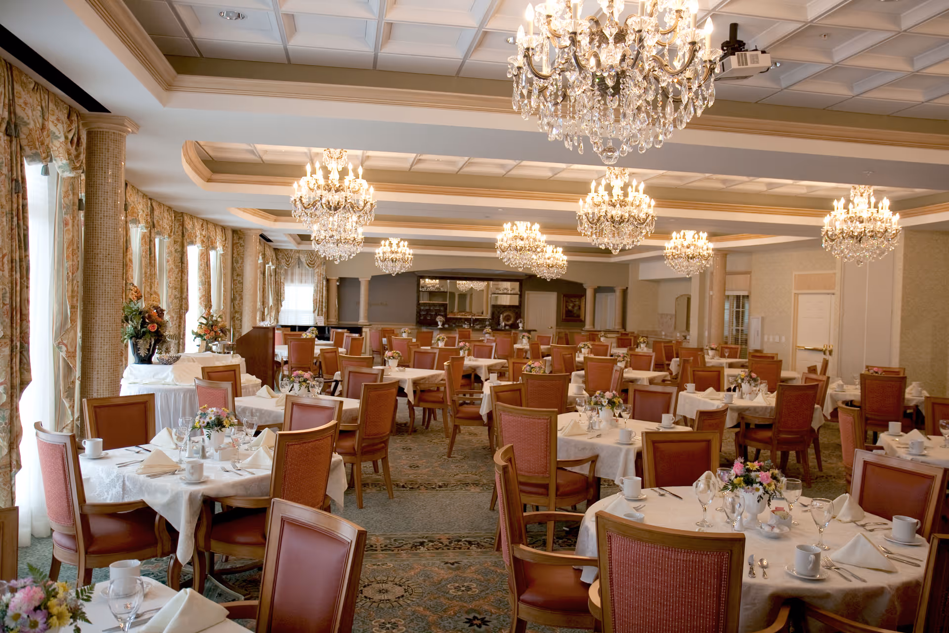 An elegant dining room with many round tables set with white linens, floral centerpieces, and crystal chandeliers.