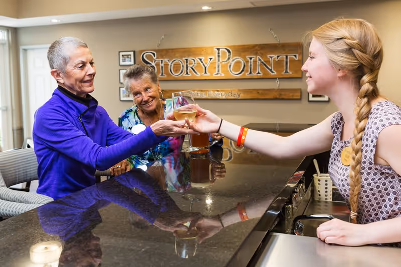 Two elderly women sitting at a counter in a communal area, one reaching out to receive a glass of white wine from a young woman with a braided hairstyle standing behind the counter. A wooden sign on the wall behind them reads 'StoryPoint'.