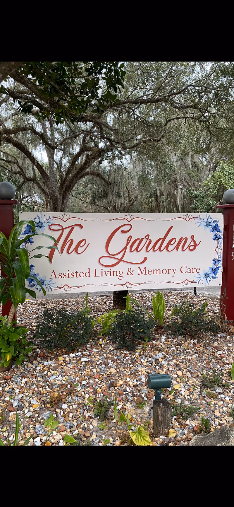 Outdoor sign for The Gardens Assisted Living & Memory Care facility surrounded by plants and trees with a gravel ground cover.