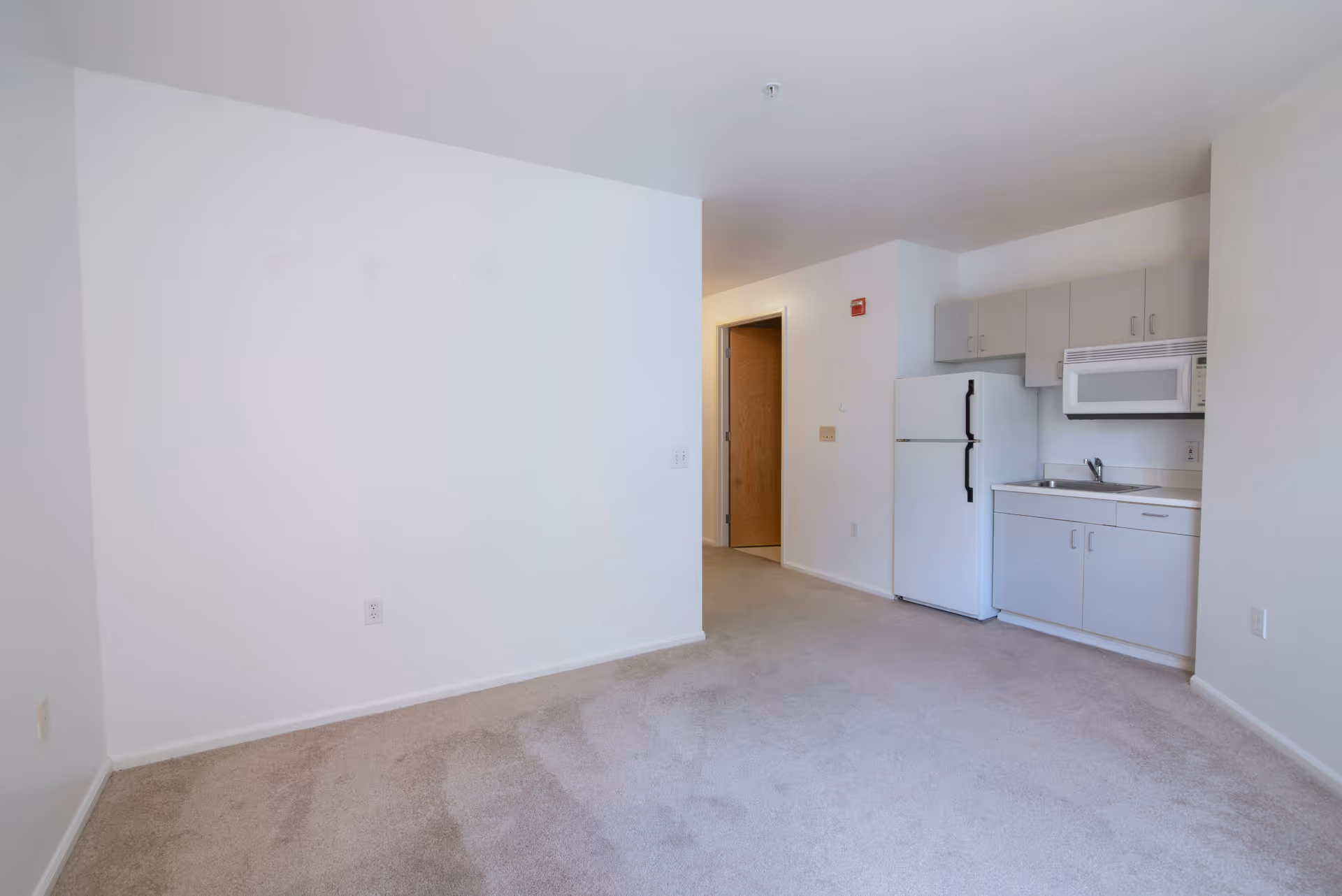 An empty room with beige carpet and white walls featuring a small kitchenette area with white cabinets, a refrigerator, a microwave, and a sink. There is an open doorway leading to another room.