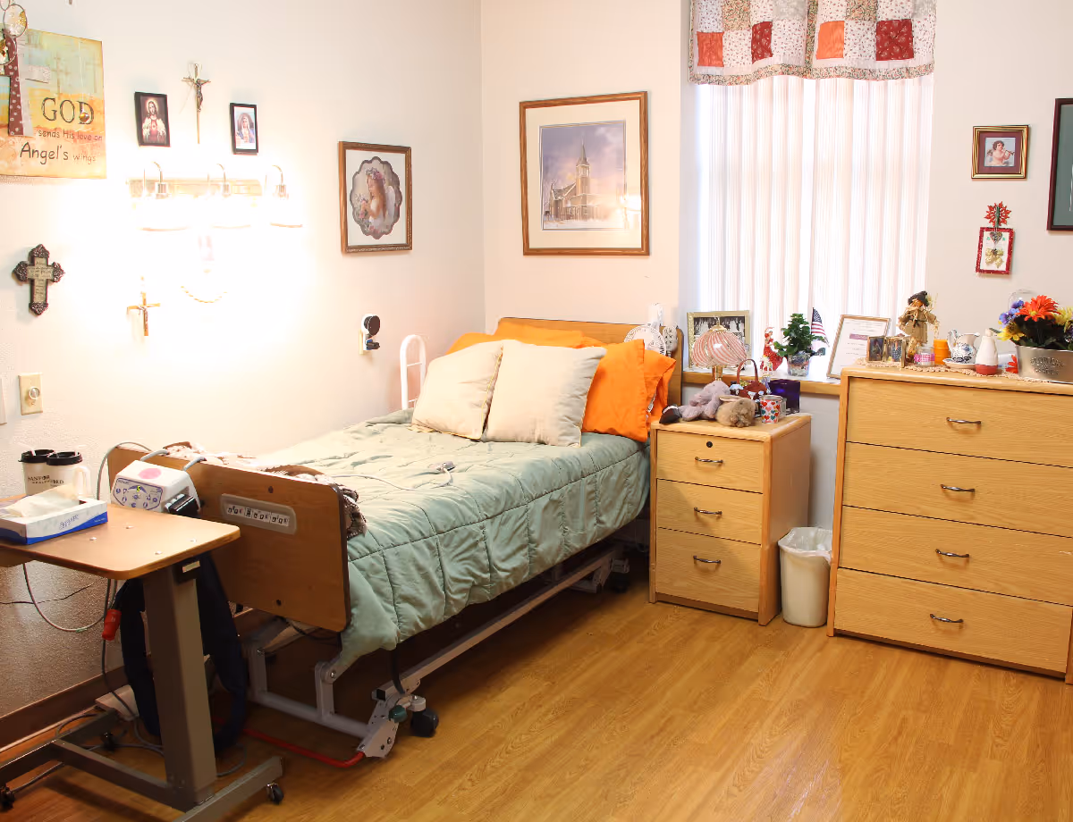 A cozy senior living bedroom with a hospital-style bed made up with green bedding and orange pillows. The room has wooden flooring, a small bedside table with stuffed animals and personal items, a larger dresser with flowers and framed pictures, and religious-themed wall decorations. A window with vertical blinds and a patchwork valance lets in natural light.