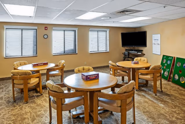 A well-lit common room with three round wooden tables, each surrounded by four cushioned chairs. Board games are placed on the tables. The room has three windows with blinds, a wall-mounted flat screen TV, and two green game boards leaning against the wall. The carpet has a beige patterned design and the walls are painted a light tan color.