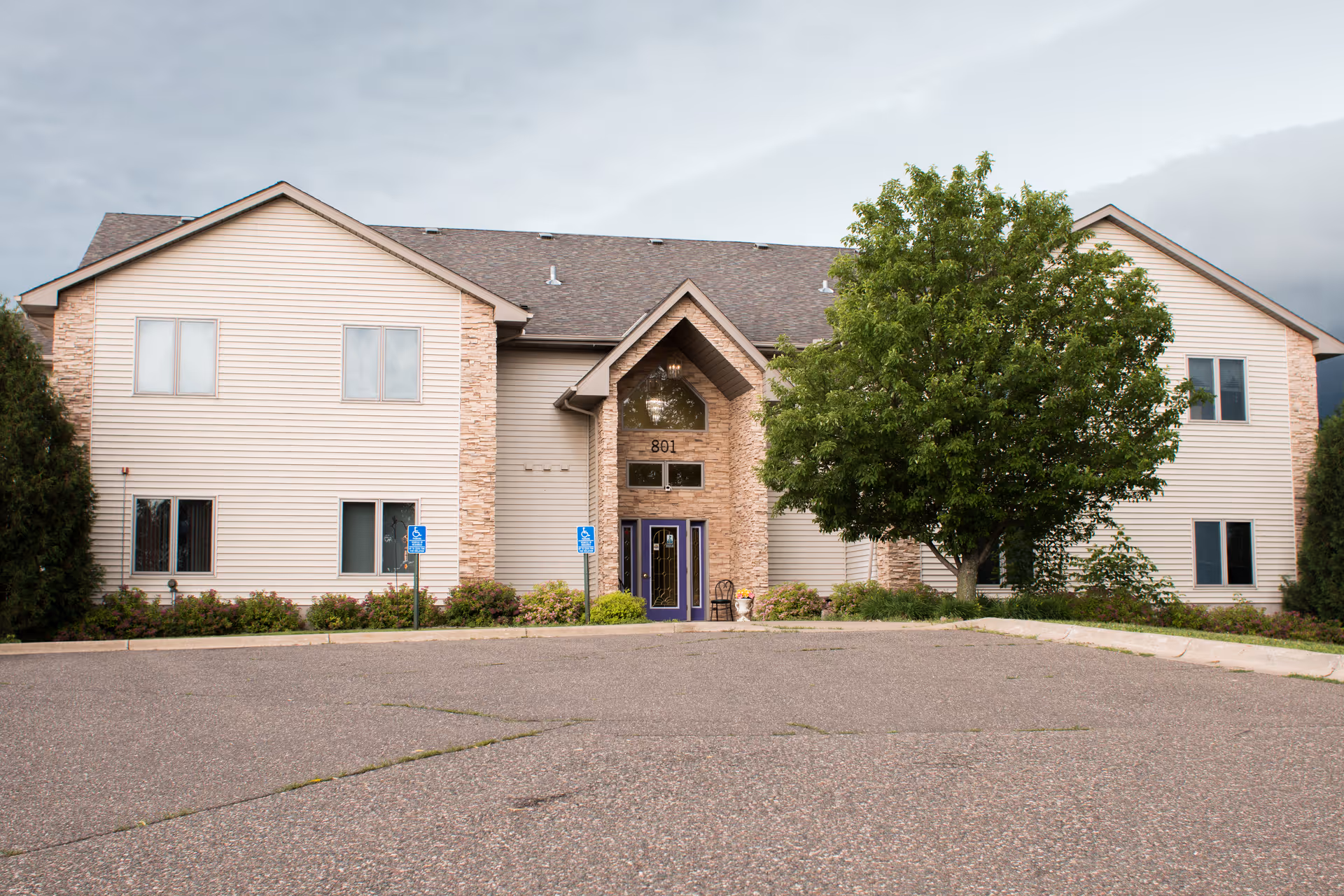 Exterior front view of a two-story senior living facility building with beige siding and stone accents. The entrance has a peaked roof and double doors painted purple. There are two handicap parking signs near the entrance and a large green tree to the right of the doorway. The sky is cloudy.