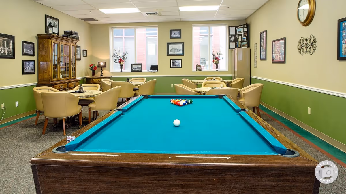 Interior room with a pool table in the foreground and several round tables with beige chairs in the background. The walls are painted green and beige, decorated with framed pictures and a clock. There is a wooden cabinet on the left side and windows letting in natural light.
