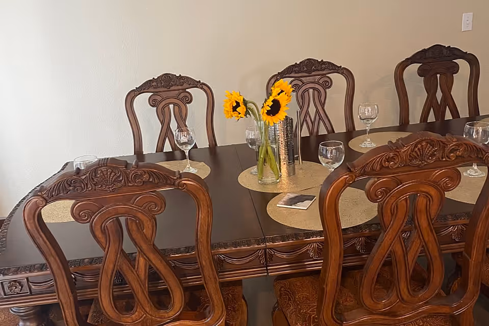 A dark wood dining table with ornate carved chairs, place settings, wine glasses and a vase of sunflowers.