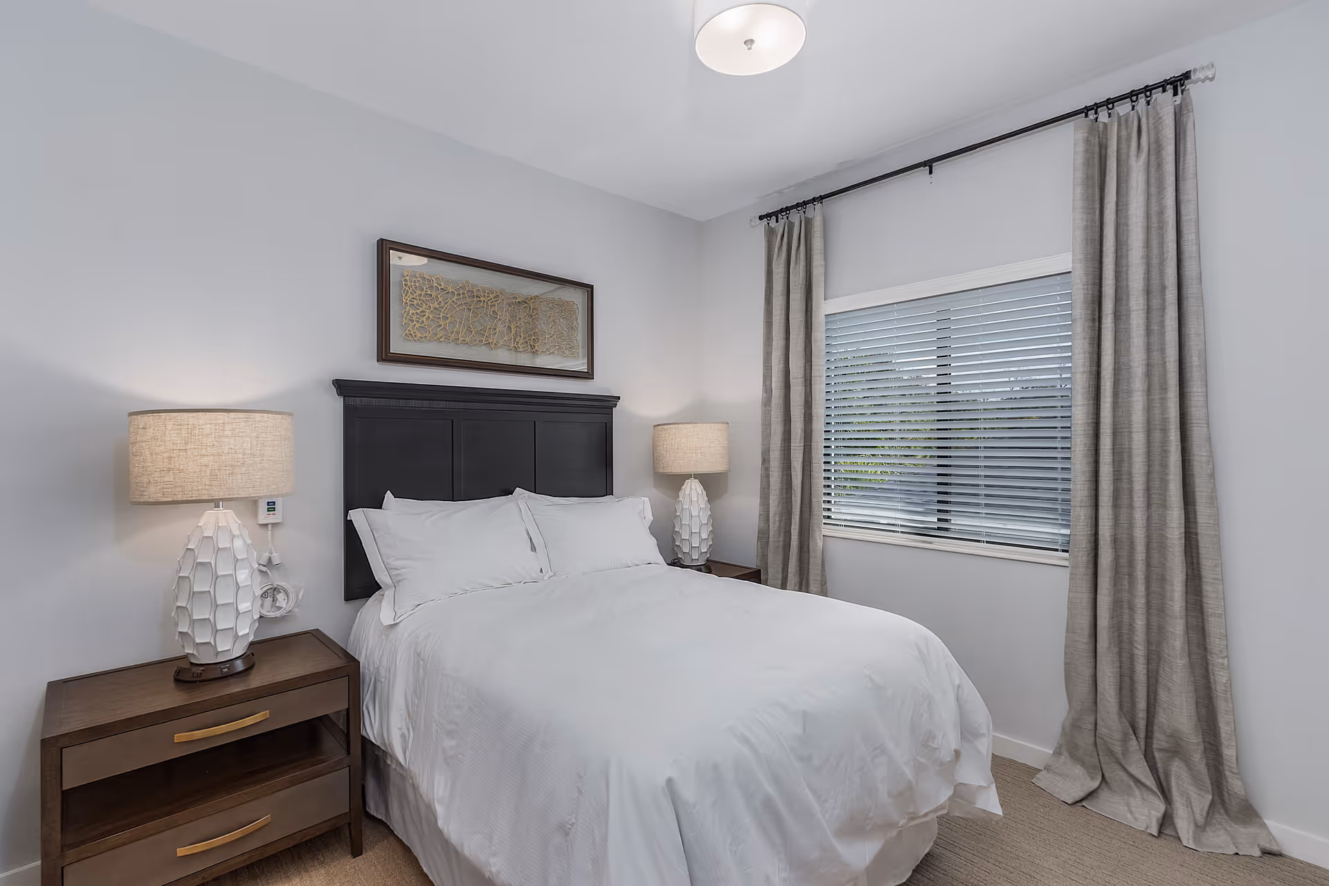 A neatly made bed with white linens and pillows in a bedroom. The bed has a dark wooden headboard and is flanked by two matching nightstands, each with a white textured lamp. There is a window with closed blinds and beige curtains on the right wall, and a framed abstract artwork hangs above the headboard. The walls are painted light gray and the room has a ceiling light fixture.