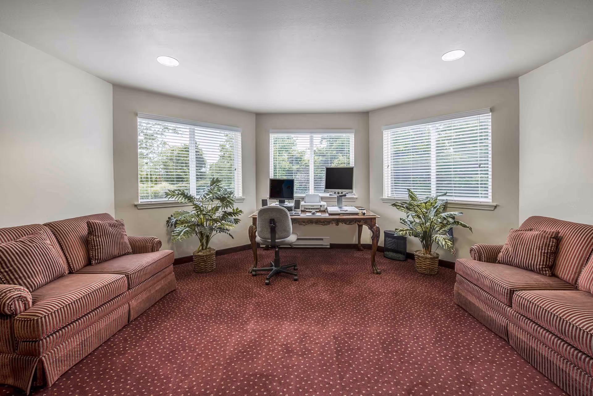 A quiet office or common room with two red and white striped sofas facing each other on a red patterned carpet. Between the sofas is a wooden desk with two computer monitors, a printer, and a gray office chair. The room has three large windows with white blinds and two potted plants on either side of the desk.