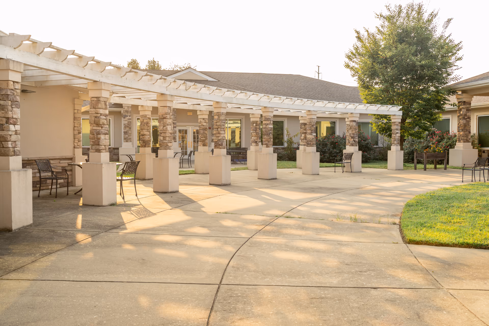 Sunlit courtyard with a pergola supported by stone columns, outdoor chairs, and a curved concrete walkway in front of the building.