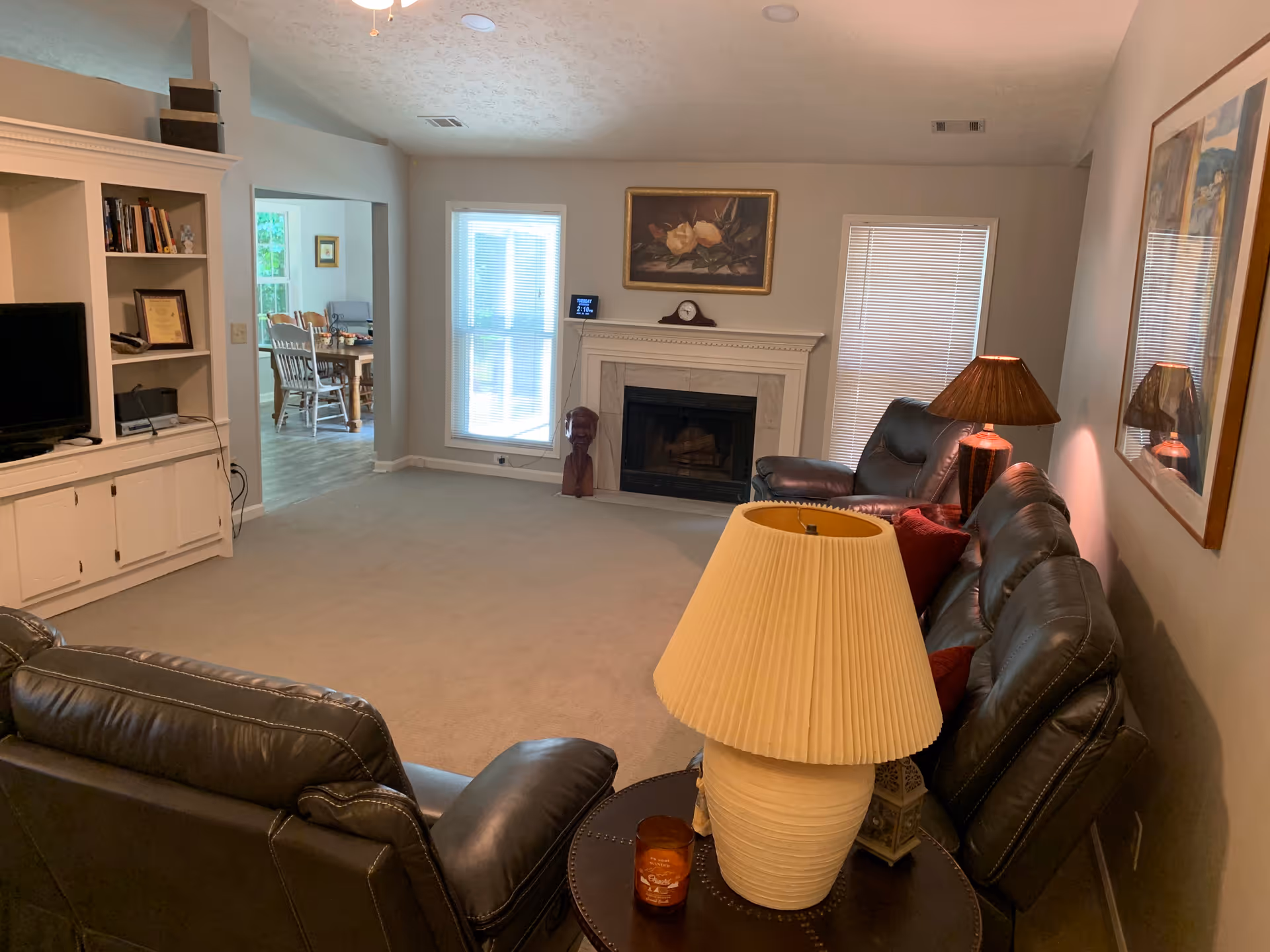 A furnished living room with leather sofas, side lamps, a fireplace, built-in shelving, and a dining area visible through a doorway.
