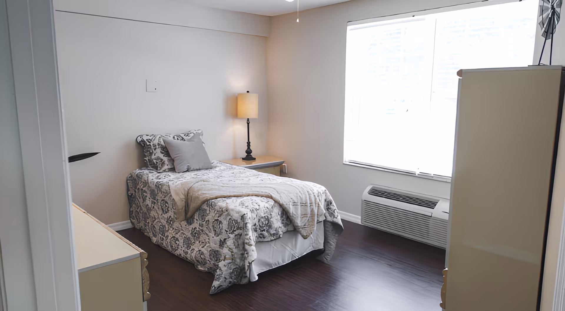 A small, neatly arranged bedroom with a single bed covered in a patterned bedspread and a gray pillow. There is a nightstand with a lamp beside the bed, a large window with blinds letting in natural light, a heating/cooling unit below the window, and a dresser on the right side of the room. The floor is dark wood, and the walls are painted white.