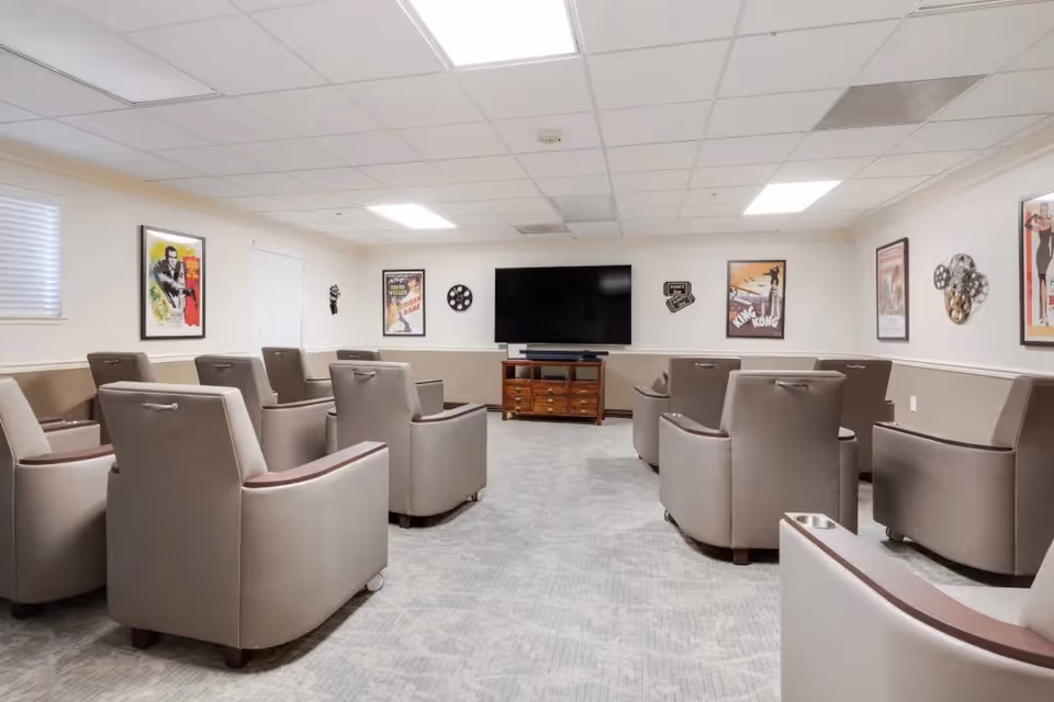 A media room with two rows of beige armchairs facing a large flat-screen TV mounted on the wall. The walls are decorated with vintage movie posters and film reel decorations. The room has a light gray carpet and a white ceiling with recessed lighting.