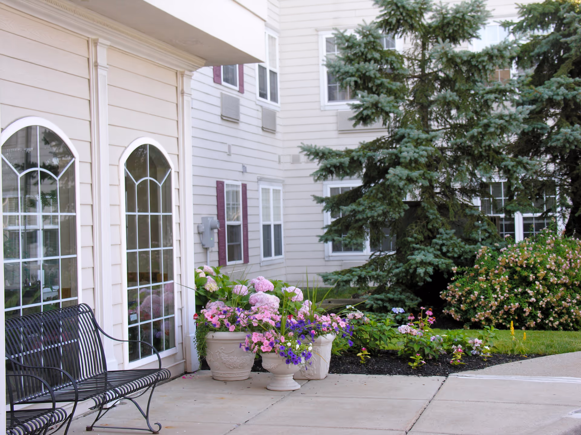 Outdoor patio area at Elderwood Assisted Living at West Seneca featuring a black metal bench, large flower pots with colorful blooming flowers, and a landscaped garden with bushes and a large evergreen tree next to the building with beige siding and multiple windows.