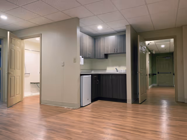 Interior view of a senior living facility showing a small kitchenette with dark wood cabinets, a mini refrigerator, and a sink. To the left, an open door reveals a bathroom with a shower and grab bars. To the right, an open door leads to a hallway with green walls and a closed door at the end. The floor is wood laminate and the ceiling has recessed lighting.