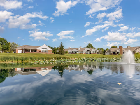 A scenic view of a senior living community with single-story buildings made of brick and siding, situated behind a pond with water lilies and a fountain. The sky is blue with scattered white clouds, and the buildings and sky are reflected in the calm water of the pond.