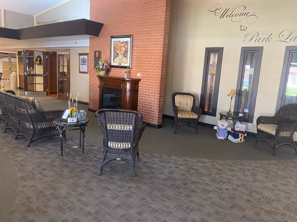 Lobby seating area with wicker chairs, small tables, a brick fireplace, and wall lettering that reads 'Welcome to Park Lane'.