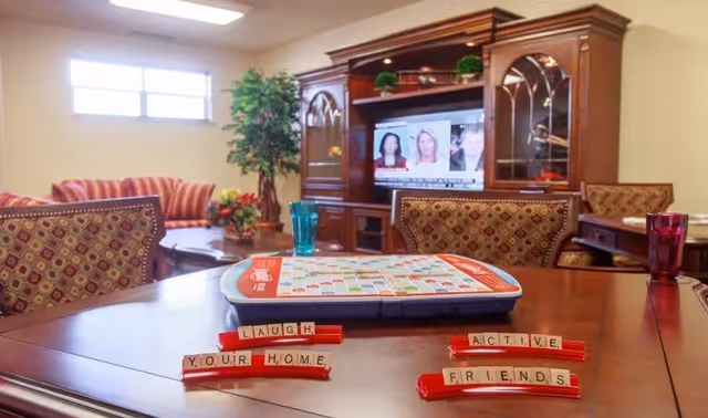 A cozy living room area with a wooden table in the foreground holding a Scrabble game with tiles spelling out 'LAUGH', 'YOUR HOME', 'ACTIVE', and 'FRIENDS'. In the background, there is a wooden entertainment center with a TV showing a news program, a striped sofa, a potted plant, and patterned chairs around the table.