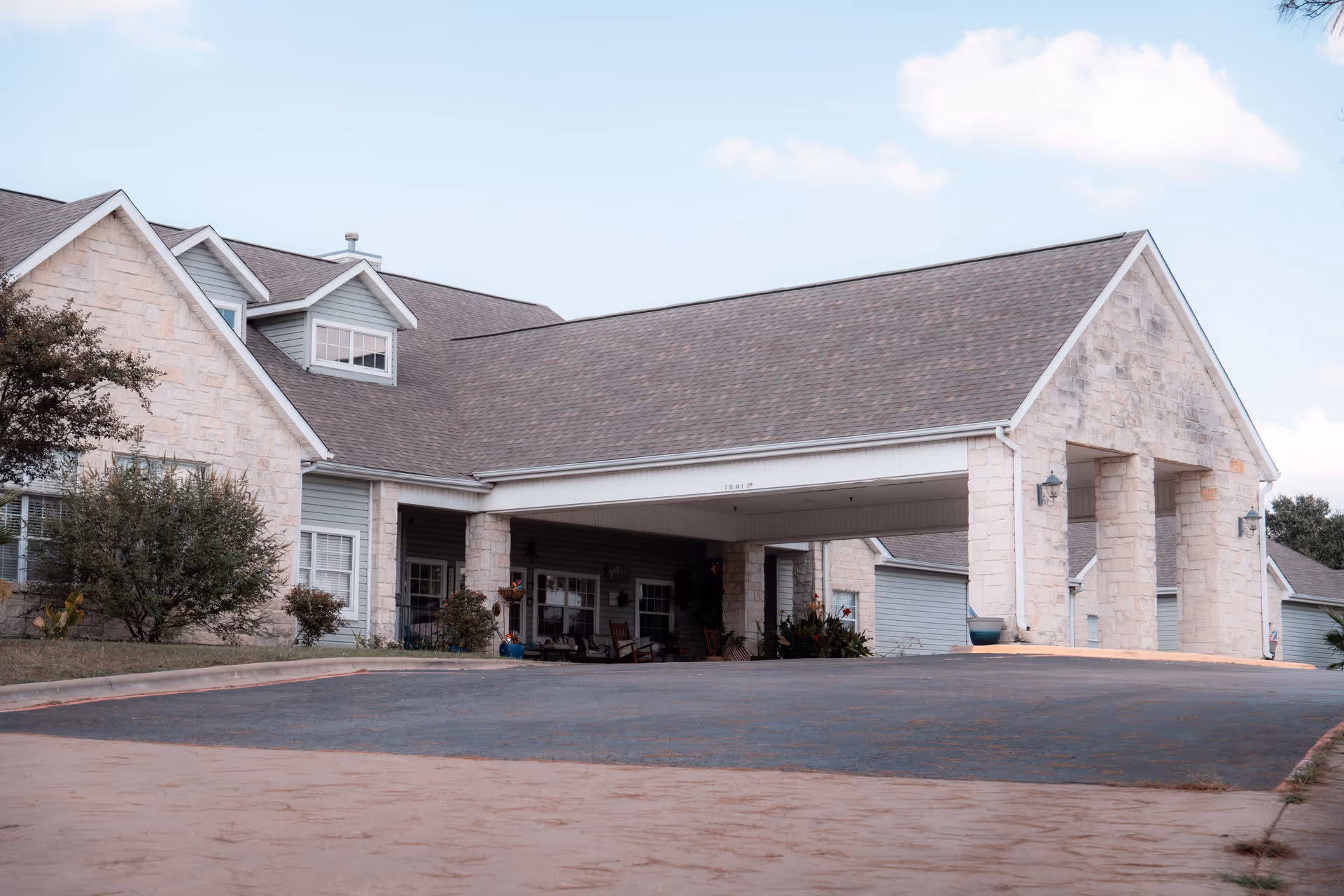Exterior view of Argent Court Assisted Living facility showing a stone and siding building with a covered entrance driveway, some bushes and plants around the building, and a partly cloudy sky.