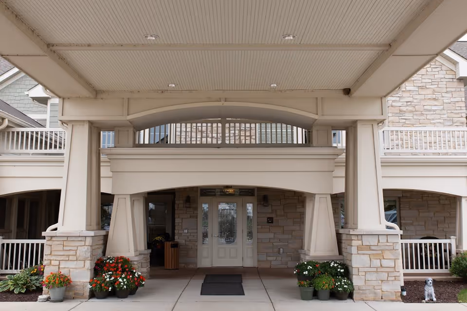 Entrance of a building with a covered porch supported by large white pillars and stone bases. There are flower pots with colorful flowers on either side of the entrance, and a small dog statue is visible on the right side near the bushes. The building has a stone facade and white railings on the upper balcony.