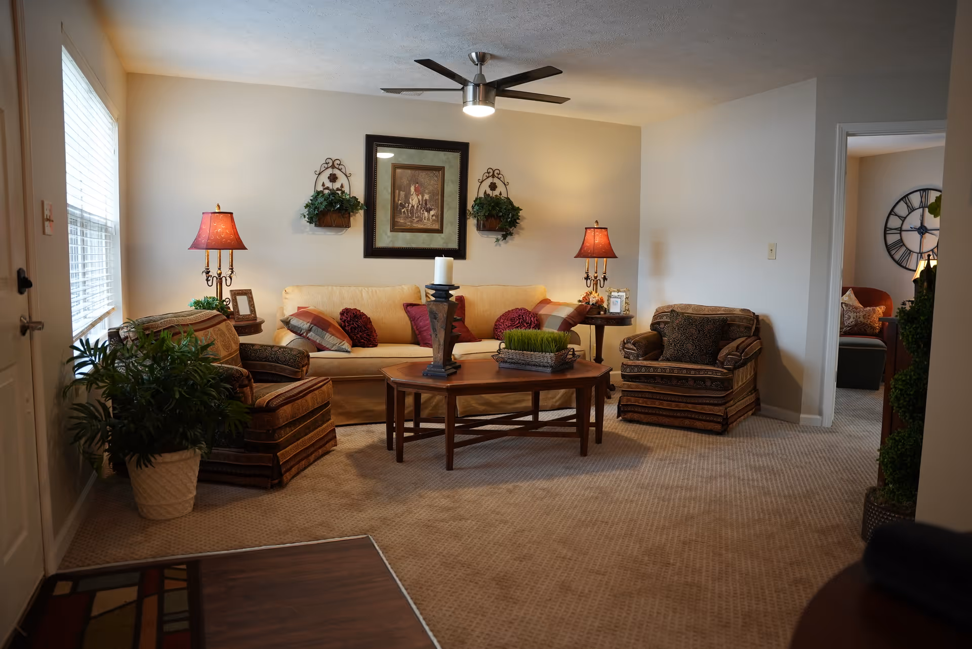 A cozy living room with a beige sofa adorned with multiple cushions, two patterned armchairs, a wooden coffee table with decorative items including a candle and a small plant, two side tables each with a lamp, wall decorations including a framed painting and two hanging plants, a ceiling fan with a light, and a large window letting in natural light.