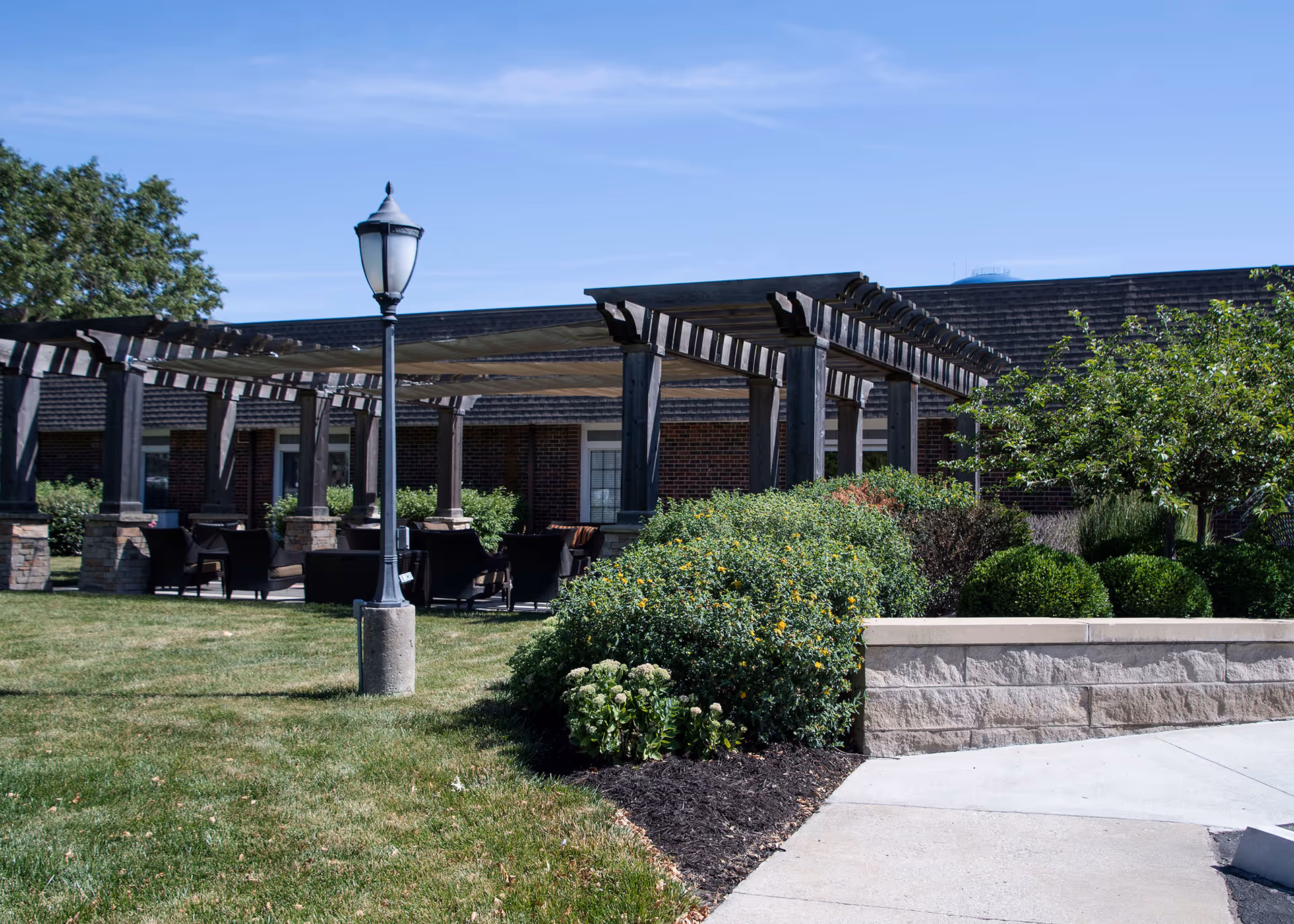 Outdoor seating area with a pergola structure, surrounded by green bushes and trees, with a lamppost in the foreground and a brick building in the background under a clear blue sky.