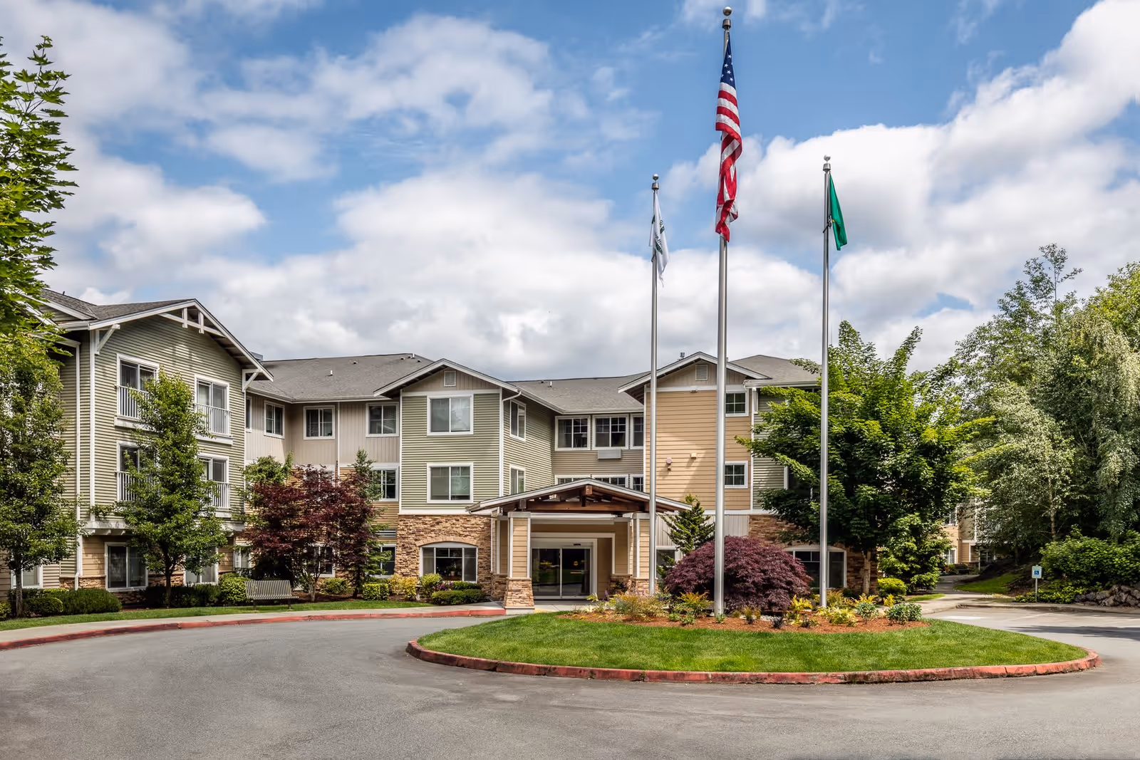 Exterior view of Woodland Terrace, a multi-story senior living facility with beige and light green siding, stone accents, and a covered entrance. Three flagpoles with flags stand in front of the building, surrounded by landscaped greenery and trees under a partly cloudy sky.