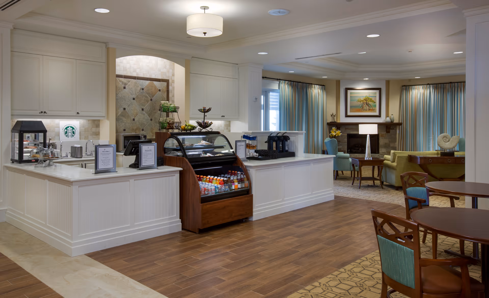 Interior view of a senior living facility cafe area with a white counter displaying a variety of beverages and snacks. Behind the counter is a small kitchen area with white cabinets and a Starbucks logo on the backsplash. To the right, there is a cozy seating area with armchairs, a fireplace, framed artwork, and large windows with curtains. Wooden tables and chairs are arranged in the foreground on a patterned carpet and wood floor.