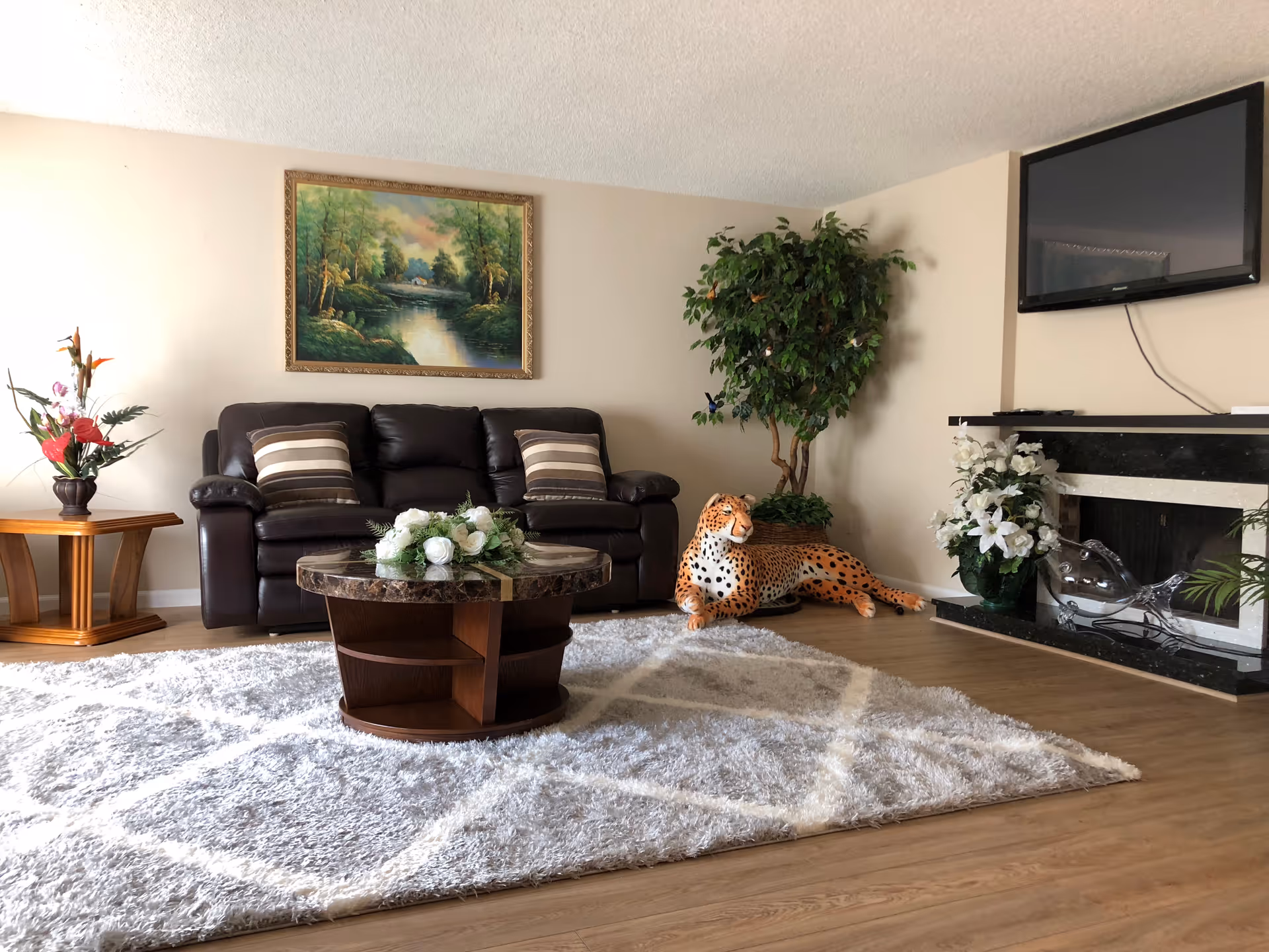 A cozy living room with a dark leather sofa, round coffee table on a plush rug, wall painting, TV above a fireplace, potted plants and a decorative leopard statue.