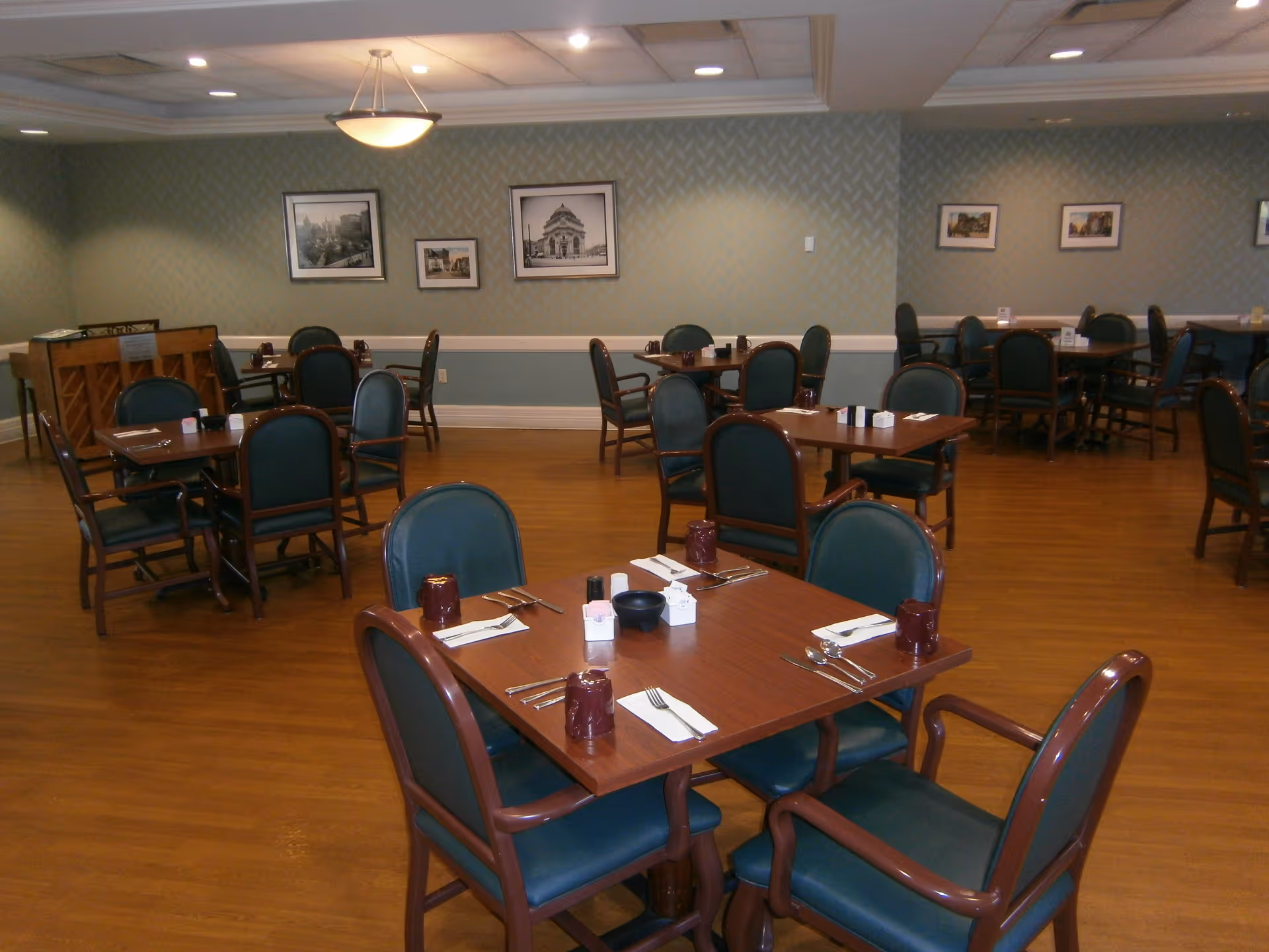 A senior living community dining room with multiple wooden tables and green cushioned chairs arranged neatly. Each table is set with utensils, napkins, and cups. The room has wooden flooring, green patterned wallpaper, framed black and white pictures on the walls, and ceiling lights providing illumination.
