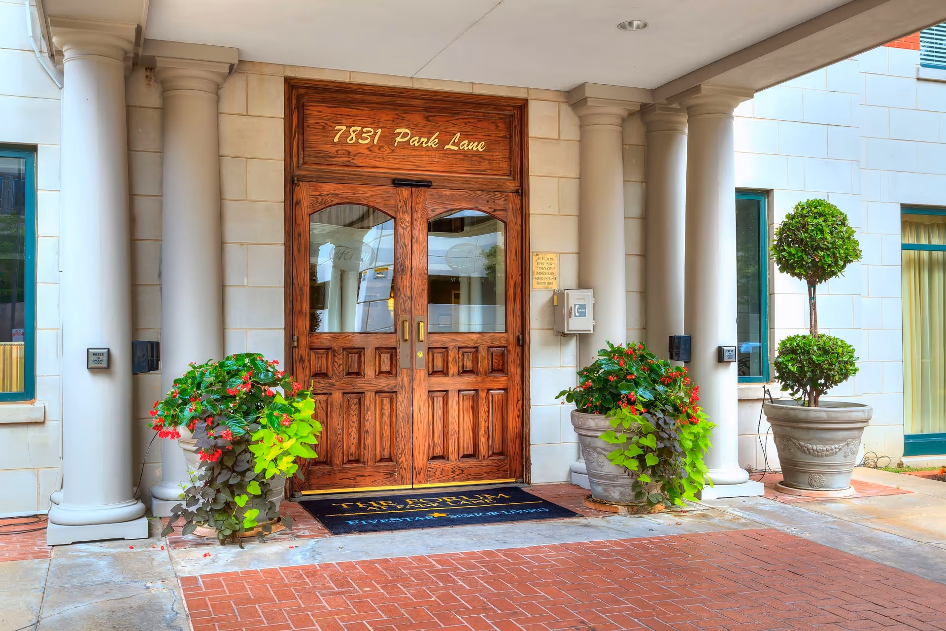 Entrance to a building with double wooden doors under the address number 7831 Park Lane, flanked by two large white columns and potted plants with green and red foliage on either side.