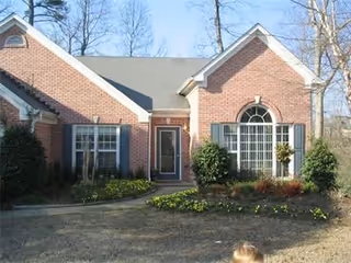 Front exterior view of a single-story brick house with a gray roof, large windows with shutters, a central front door, and a small garden with shrubs and flowers in front.