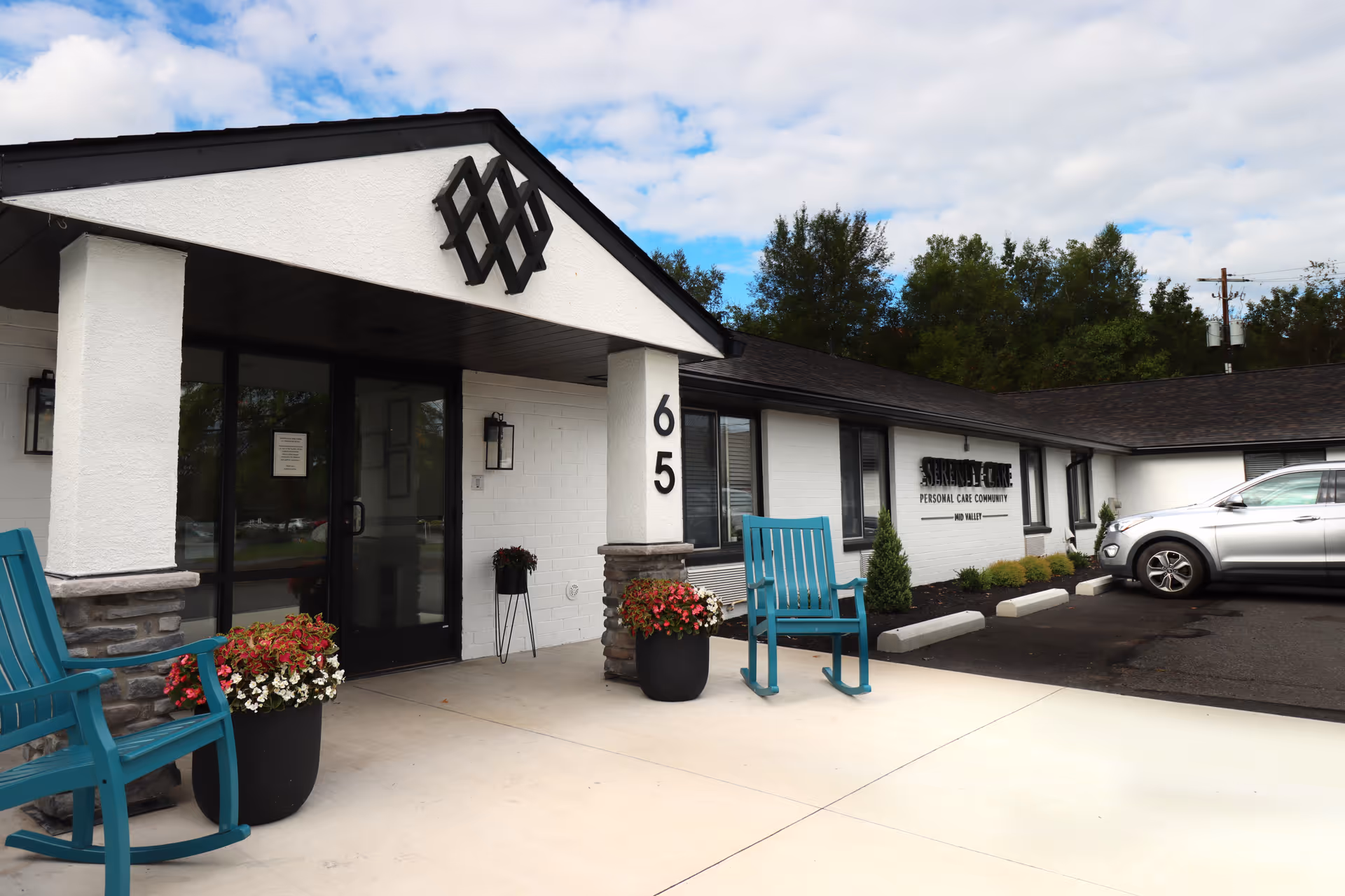 Exterior view of Serenity Care Mid Valley facility showing the entrance with a covered porch, two blue rocking chairs, flower pots, and a parked silver car. The building is white with black trim and has the number 65 on a pillar near the door.