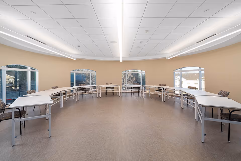 Large circular multipurpose room with folding tables and chairs arranged around the perimeter beneath recessed ceiling lights and arched windows.