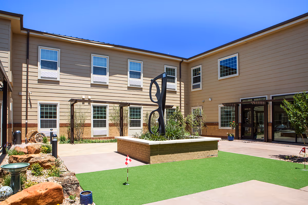 Sunny central courtyard of The Legacy at South Plains with a modern sculpture, small putting green, and a two-story beige building with entrance doors.