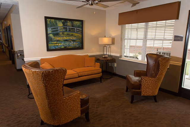 A cozy seating area in a senior living facility with a brown sofa, two patterned armchairs, a side table with a lamp, a large window with blinds, and a painting of a bridge over a pond on the wall.