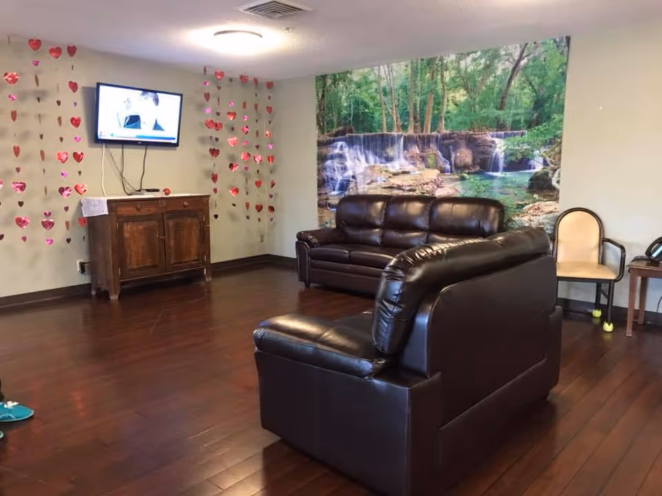 A cozy living room area with two dark brown leather sofas facing a wall-mounted TV. The wall behind the sofas features a large mural of a forest waterfall scene. Red heart-shaped decorations hang on the adjacent wall near a wooden cabinet beneath the TV. There is also a beige chair with tennis balls on its legs and a small side table with a phone.