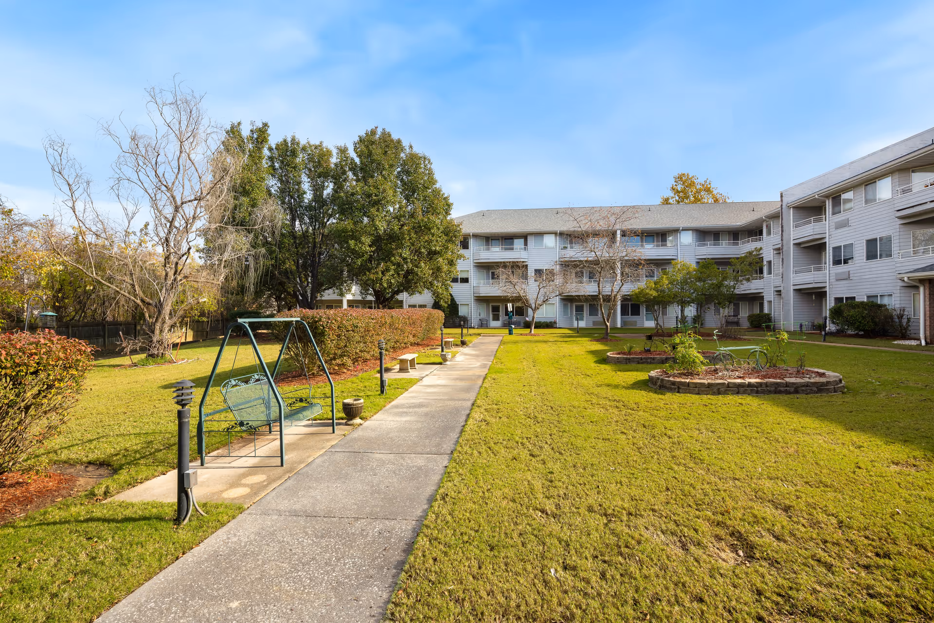 Outdoor courtyard area of Jackson Meadow - A Provincial Senior Living Community, featuring a concrete walkway, green lawn, garden beds, benches, a swing, and a three-story residential building in the background under a blue sky.