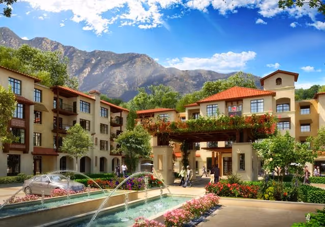 Sunny courtyard with a fountain in front of a Mediterranean-style multi-story senior living building set against mountains.
