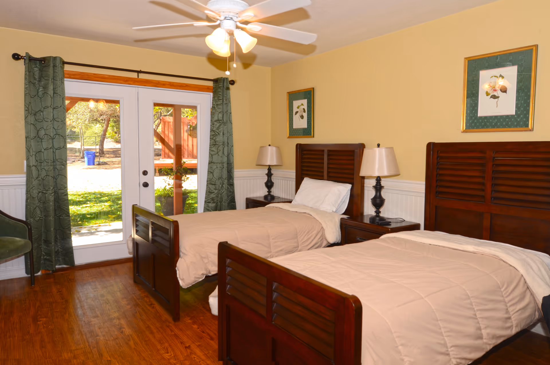 A bedroom with two single wooden beds, each with beige bedding and white pillows. Between the beds is a nightstand with a lamp. The room has yellow walls with white wainscoting, two framed botanical prints, a ceiling fan with lights, and a glass door leading outside with green curtains on either side. There is also a green chair in the corner.