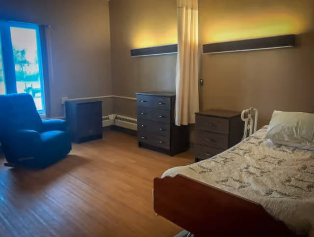 A senior living facility bedroom with a hospital-style bed covered with a patterned blanket, a pillow, and a bed rail. The room has wooden flooring, a blue recliner chair near a window, and three dark wooden dressers. A curtain hangs from a ceiling track, and a wall-mounted light fixture provides warm lighting.