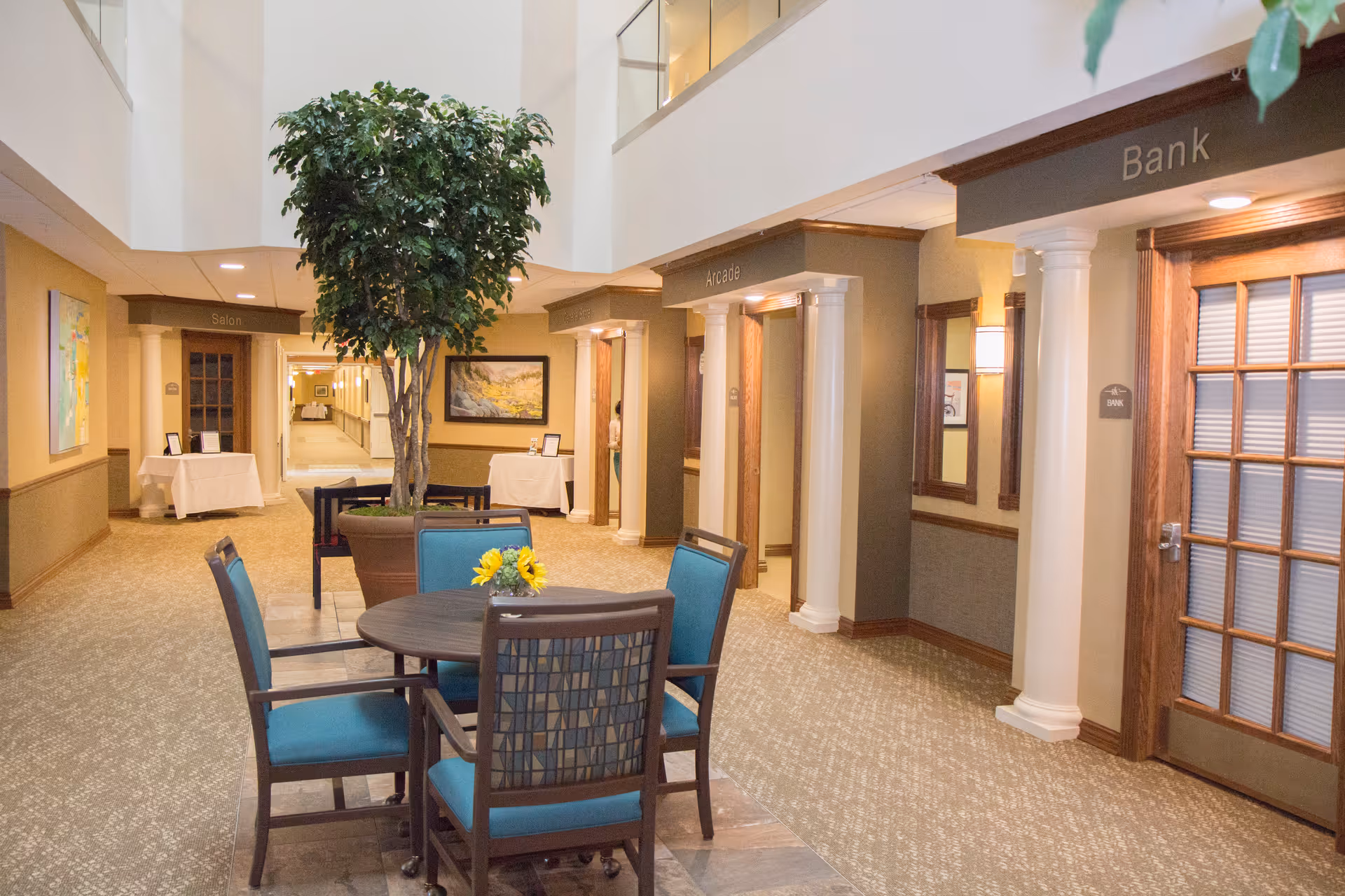 Interior hallway of a senior living facility with a round table and four chairs with blue cushions in the foreground. A large potted tree is placed near the table. The hallway has several doorways labeled Salon, Arcade, and Bank. The walls are decorated with framed artwork and the area is well-lit with ceiling lights.