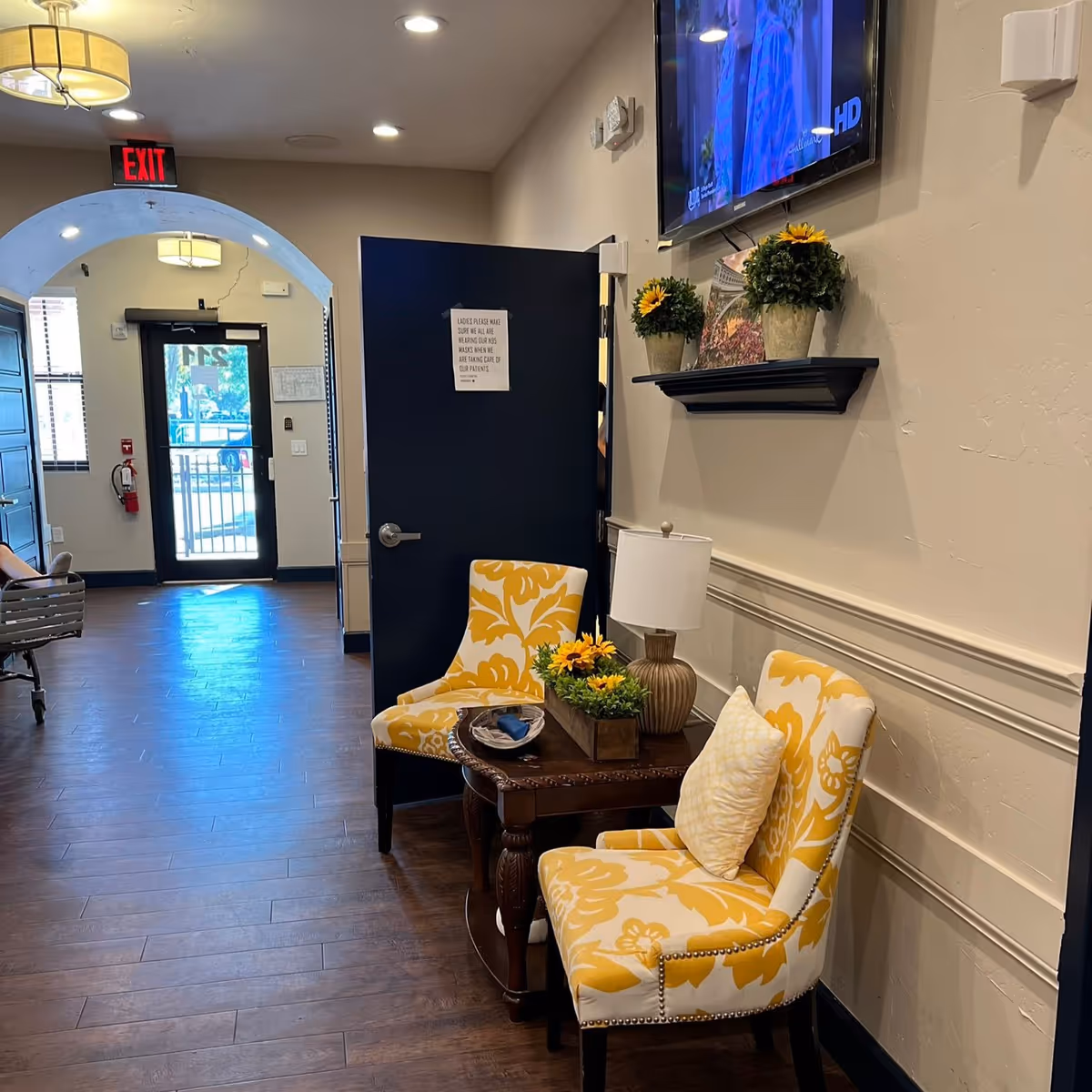 Interior view of a hallway in an assisted living facility featuring two yellow and white patterned chairs with a small wooden table between them. The table holds a lamp and a flower arrangement. Above the chairs, a wall-mounted shelf displays two flower pots and a framed picture. A flat-screen TV is mounted on the wall above the shelf. The hallway has wooden flooring, a dark blue door with a sign, and an exit door with a red exit sign above it.
