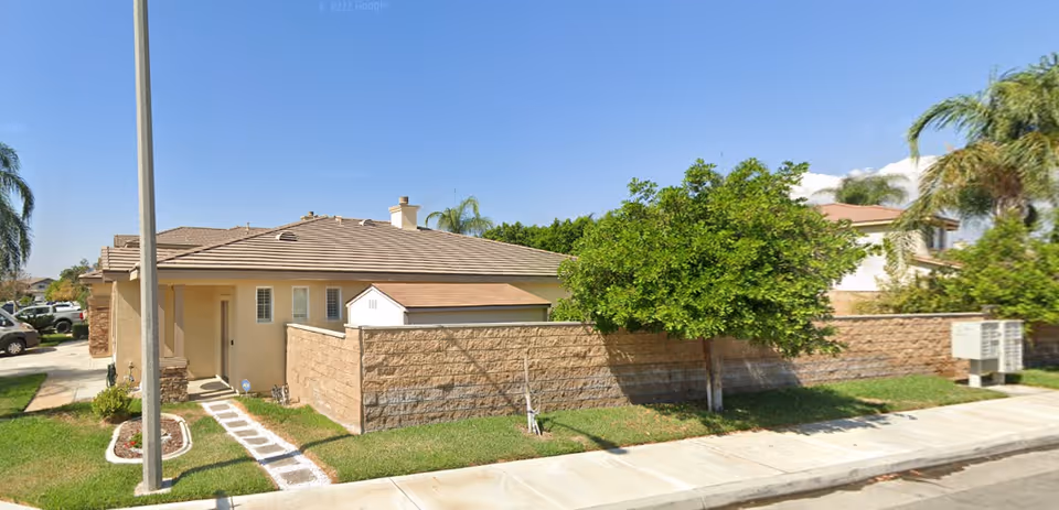 Exterior view of a single-story residential building with a tiled roof, beige walls, and a small enclosed front yard with a tree and grass. A sidewalk and street are visible in the foreground under a clear blue sky.