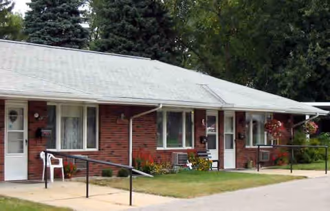Single-story brick building with white doors and windows, surrounded by greenery and trees. There are white plastic chairs and flower pots near the entrances, with a paved walkway and handrails leading to the doors.