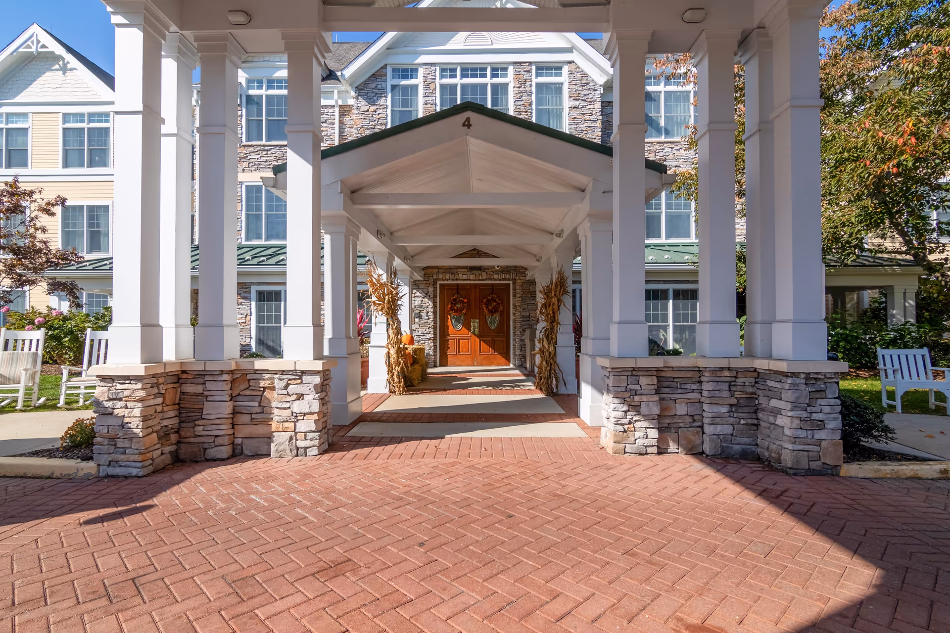Covered entrance walkway leading to double wooden doors decorated with fall wreaths, flanked by stone pillars and white columns, with a brick-paved driveway and a multi-story building in the background.