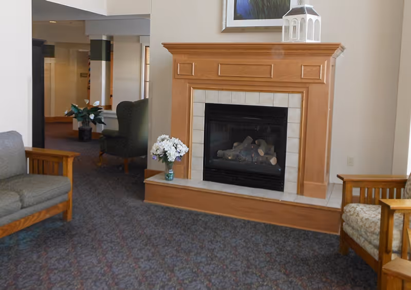 A cozy living area with a wooden fireplace surrounded by beige tiles. There are two wooden armchairs with cushions on either side of the fireplace, a gray cushioned sofa on the left, and a green armchair in the background near a potted plant. A small vase with white flowers is placed on the hearth, and a decorative lantern sits on top of the fireplace mantel.