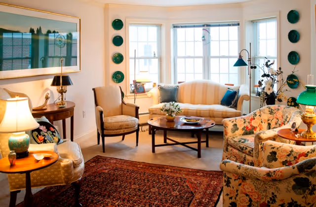 A cozy living room with a floral armchair, a striped sofa, two beige armchairs, a wooden coffee table with a small plant, and several lamps providing warm lighting. The room has a large window with white blinds, decorative plates on the walls, and a patterned area rug on the floor.