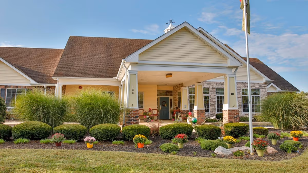 Front entrance of a single-story senior living facility with a covered portico, landscaped shrubs and potted flowers and a flagpole.