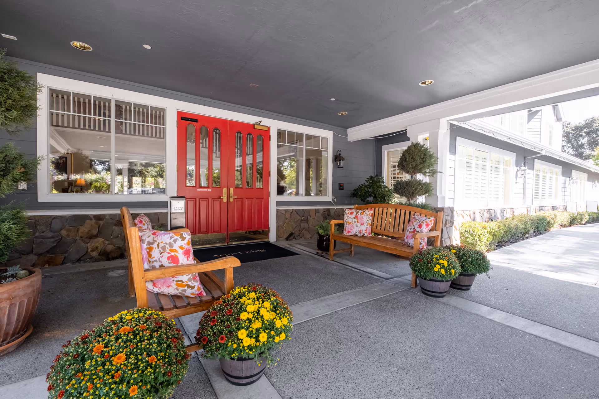 Entrance area of Aegis Living Pleasant Hill featuring a covered porch with two wooden benches adorned with floral cushions. There are potted yellow and orange flowers in front of the benches. The building has gray siding with stone accents and large windows. The entrance door is bright red with glass panels.