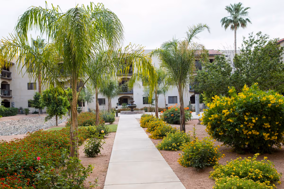 A paved walkway lined with palm trees and various flowering bushes leading to a multi-story building with balconies. The garden area features green shrubs, yellow and orange flowers, and a central fountain in front of the building under a cloudy sky.