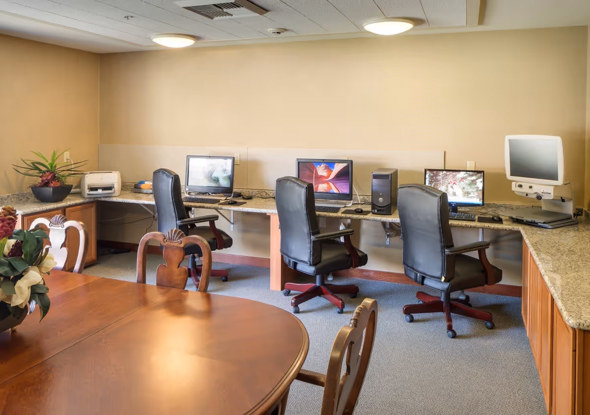 A computer room with three black office chairs in front of a long granite countertop holding three desktop computers and a printer. In the foreground, there is a wooden table with wooden chairs and a flower arrangement. The room has beige walls and carpeted floor.